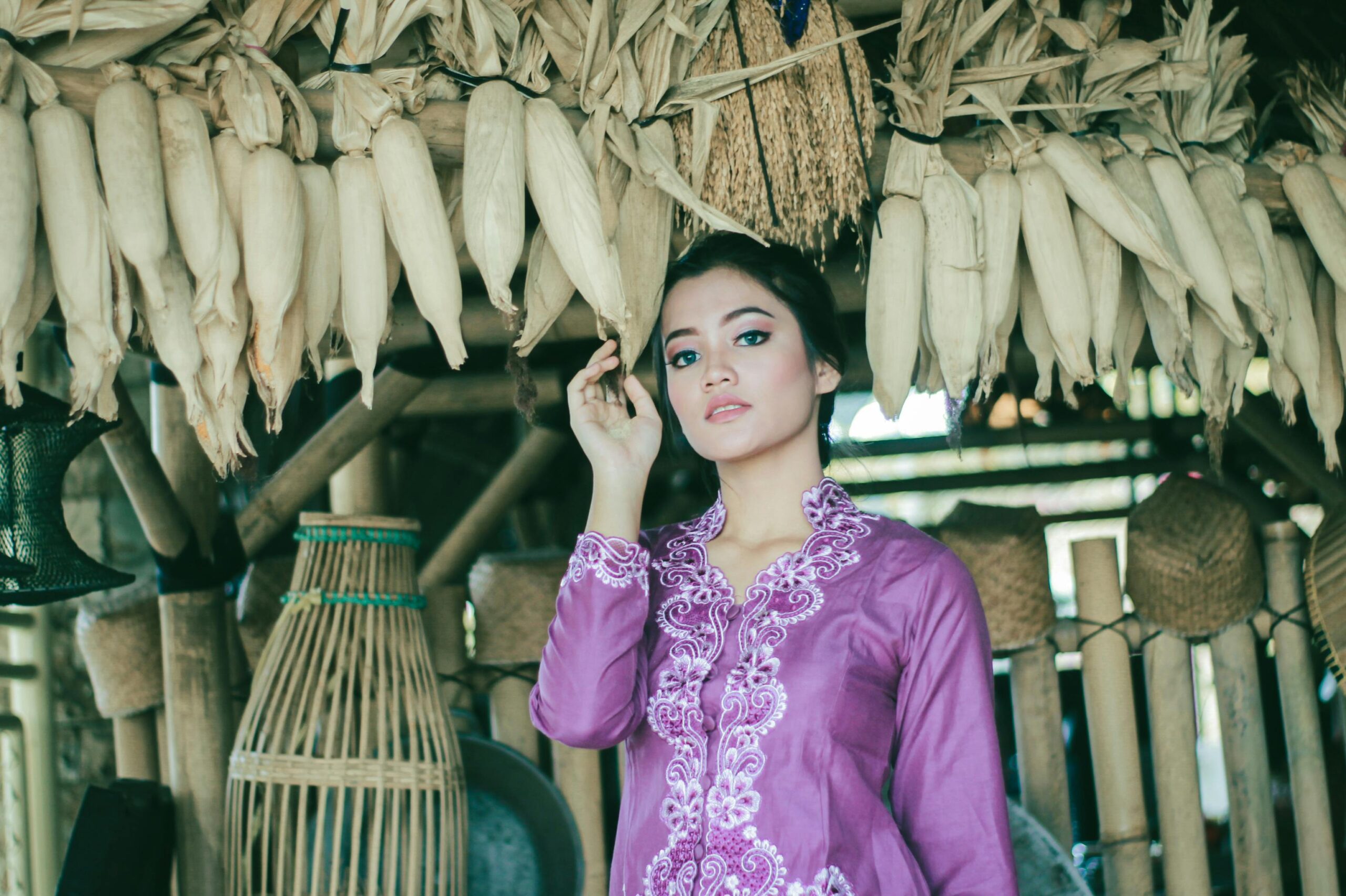 A young woman in a traditional purple dress poses indoors next to hanging corn.